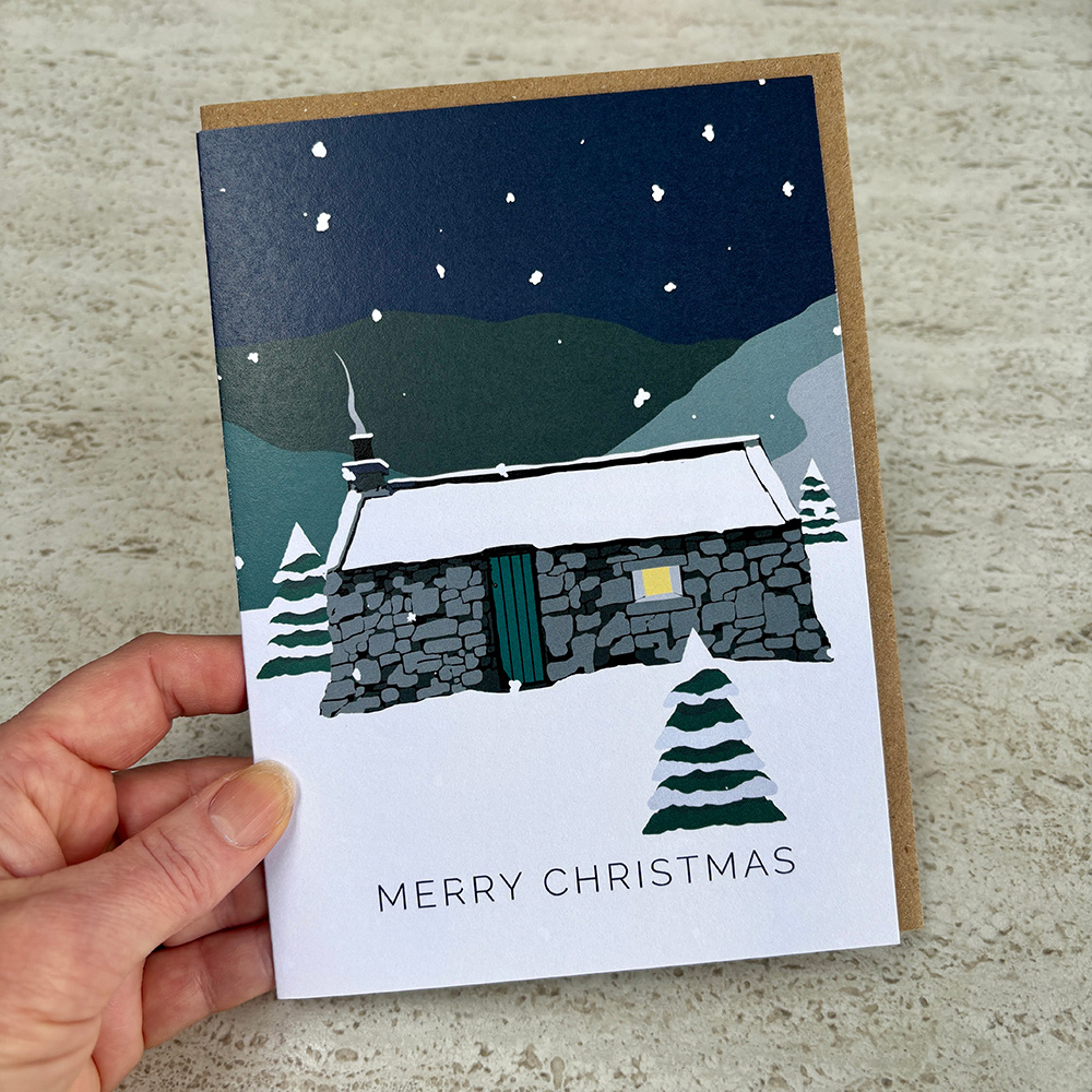 A hand holding a Christmas card of a snowy Scottish scene with a stone bothy. The window is lit with a warm yellow light and smoke is coming from the chimney.